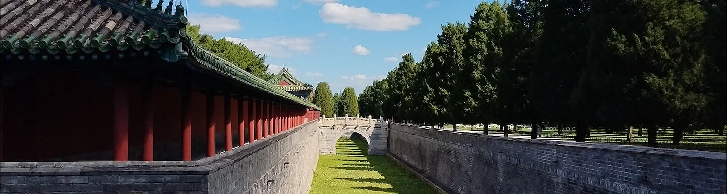 Traditional Chinese red-walled building beside a grassy canal with a small stone bridge and trees.