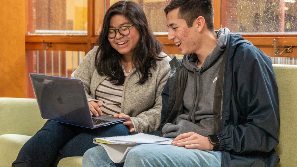 Two students on a couch pointing to a computer and smiling.
