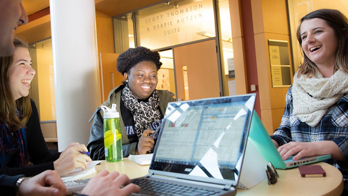 Three students sitting together at a table, working on computers and smiling while collaborating.