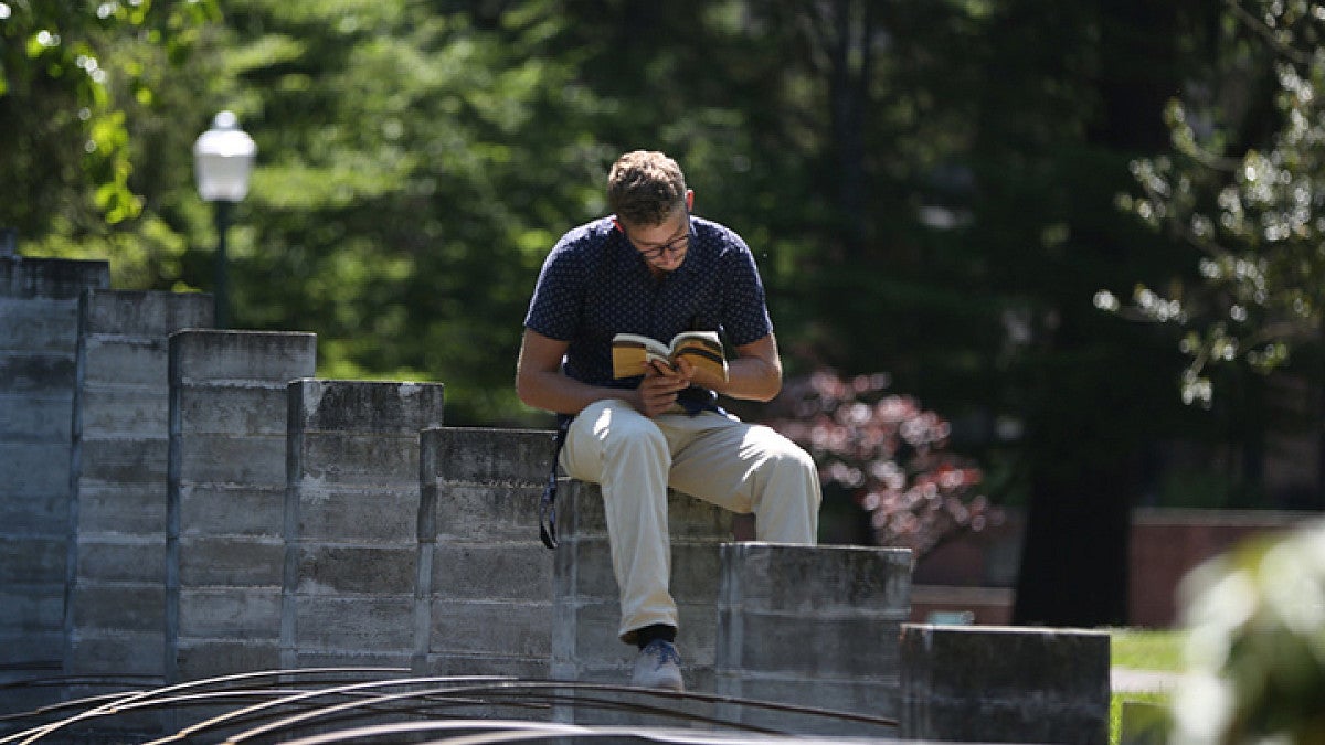A student reading a book on a college campus, sitting outdoors in a relaxed setting.