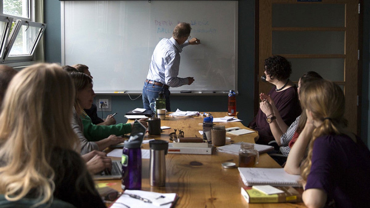 Students seated in a classroom attentively watching a professor write on the chalkboard.