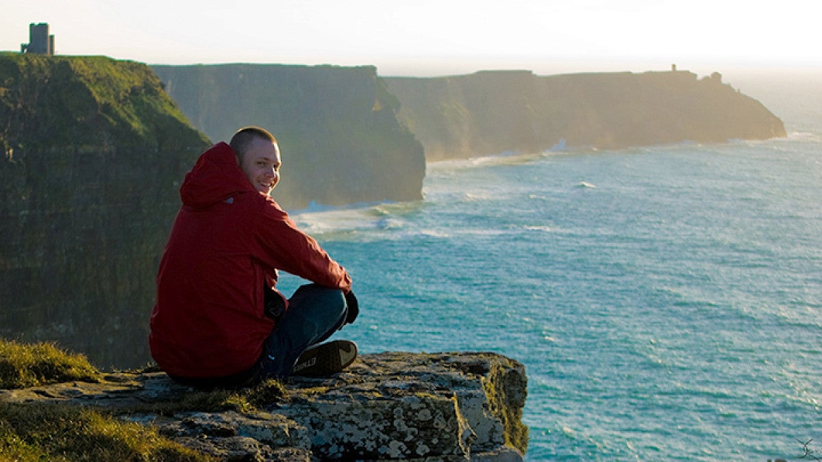 Student smiling while sitting on a cliffside overlooking the ocean on a sunny day.