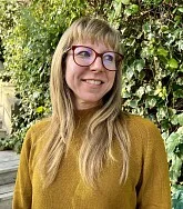photo of woman with long hair and glasses standing in front of greenery