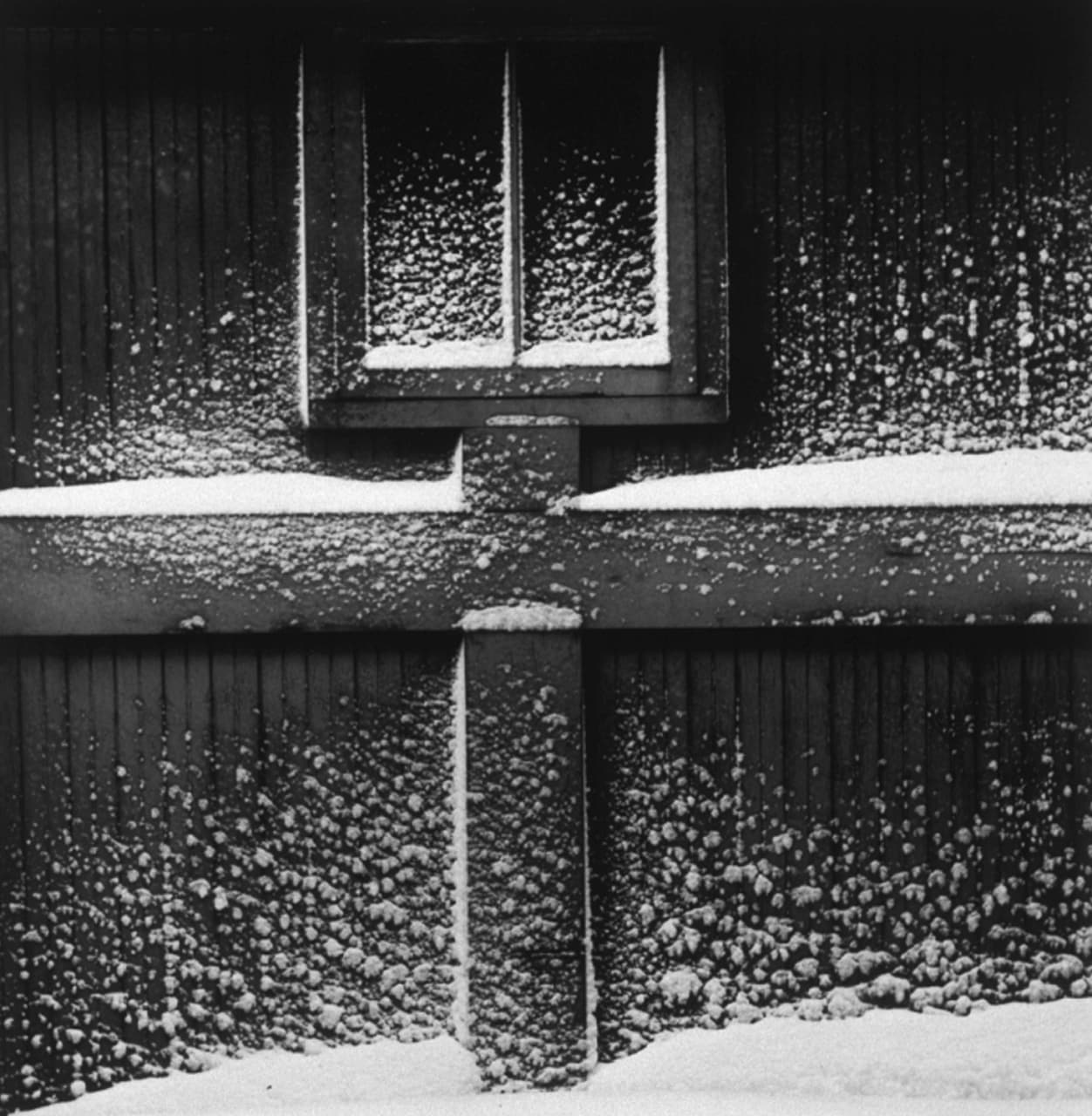 A black and white photo of a wall with a cross beam and window. The beam has snow piled on top and there's snow on the ground at the base of the wall.