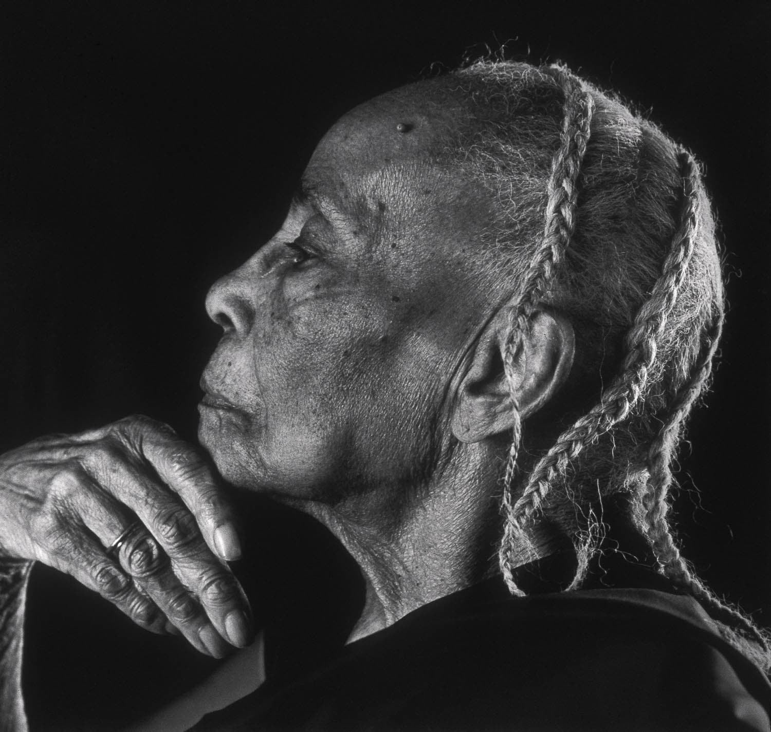 Black and white portrait of a older Black woman viewed from the left side. She is resting her chin on her hand and has multiple braids in her hair.