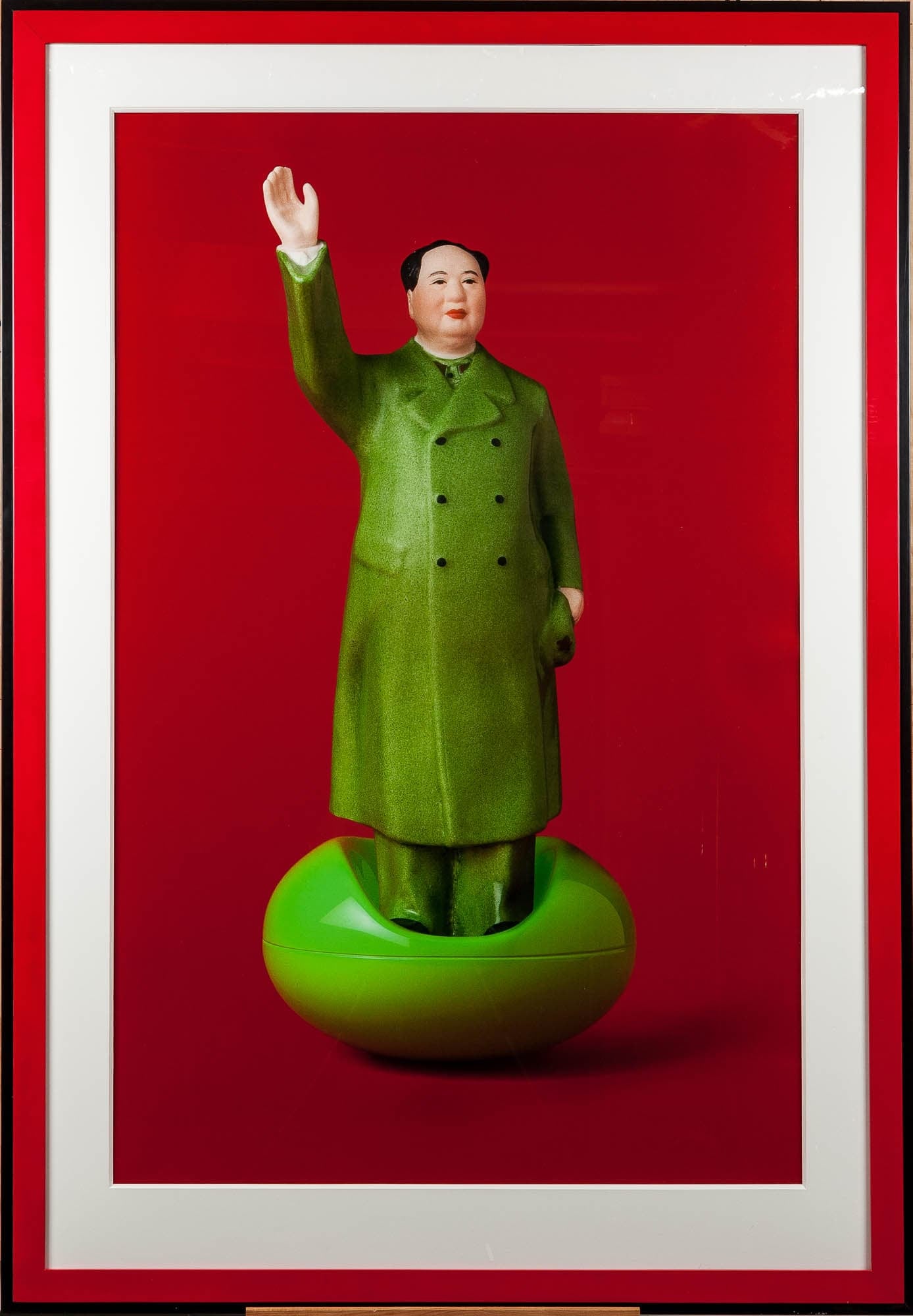 A photograph of a porcelain statue of Mao Zedong standing on a green base, set against a bright red background. The statue is in a traditional pose, with one arm raised in a gesture of greeting or command.