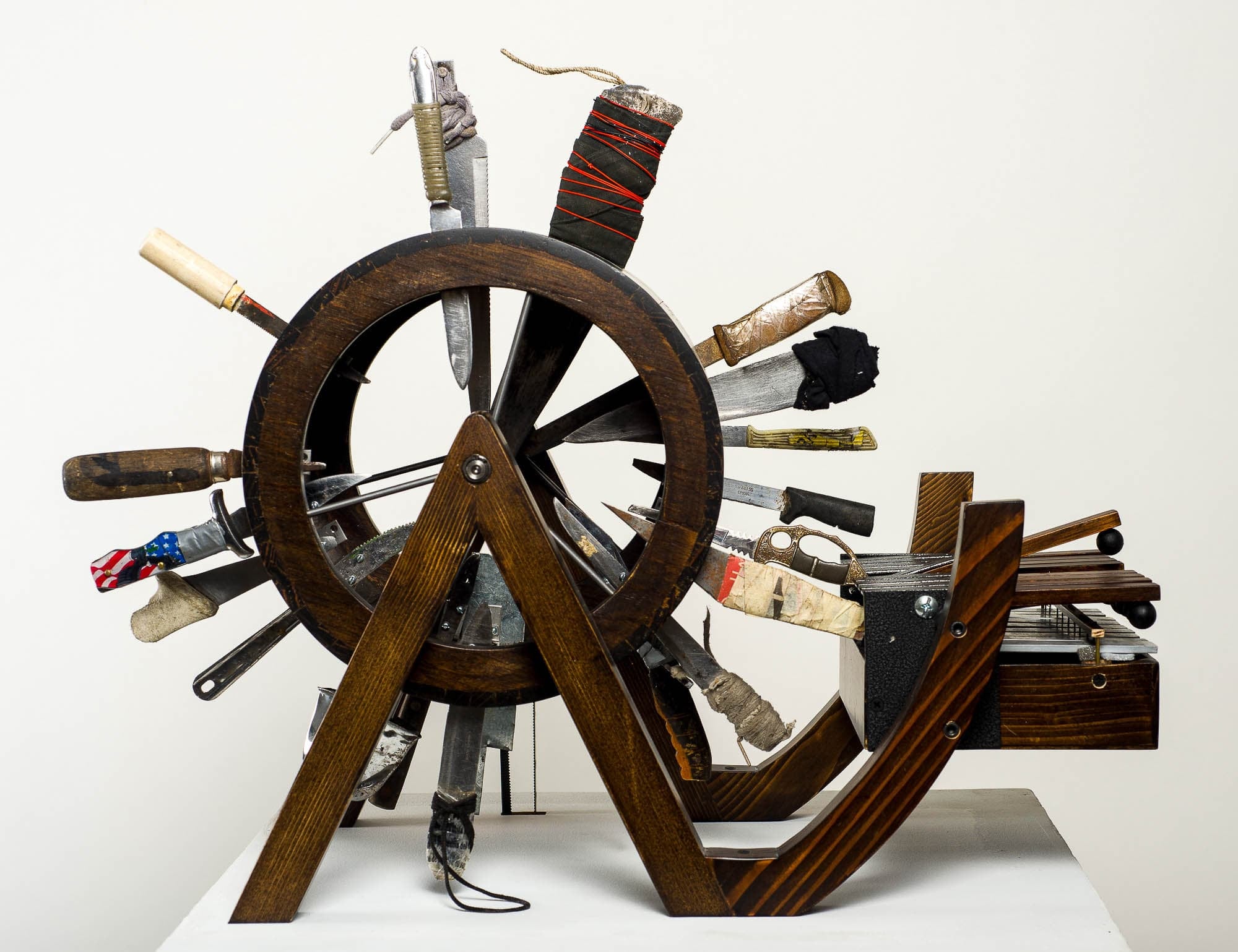 A wooden wheel with various knives and tools inserted around its perimeter, displayed against a plain background. The diverse collection of implements suggests a theme of utility and craftsmanship.