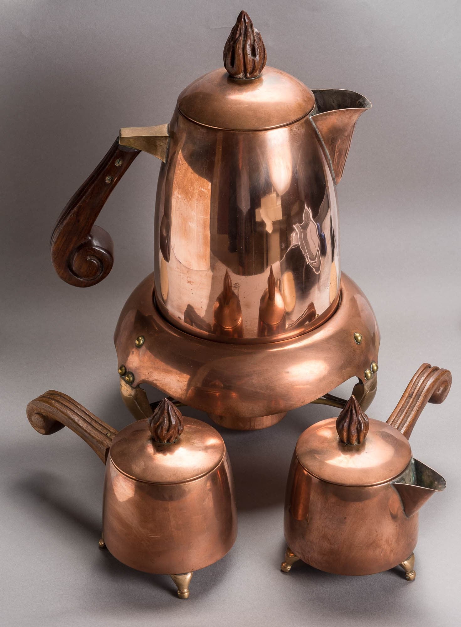 A set of copper kitchenware, including a kettle and two smaller pots, with wooden handles and decorative finials. The polished surfaces reflect light, highlighting their craftsmanship.