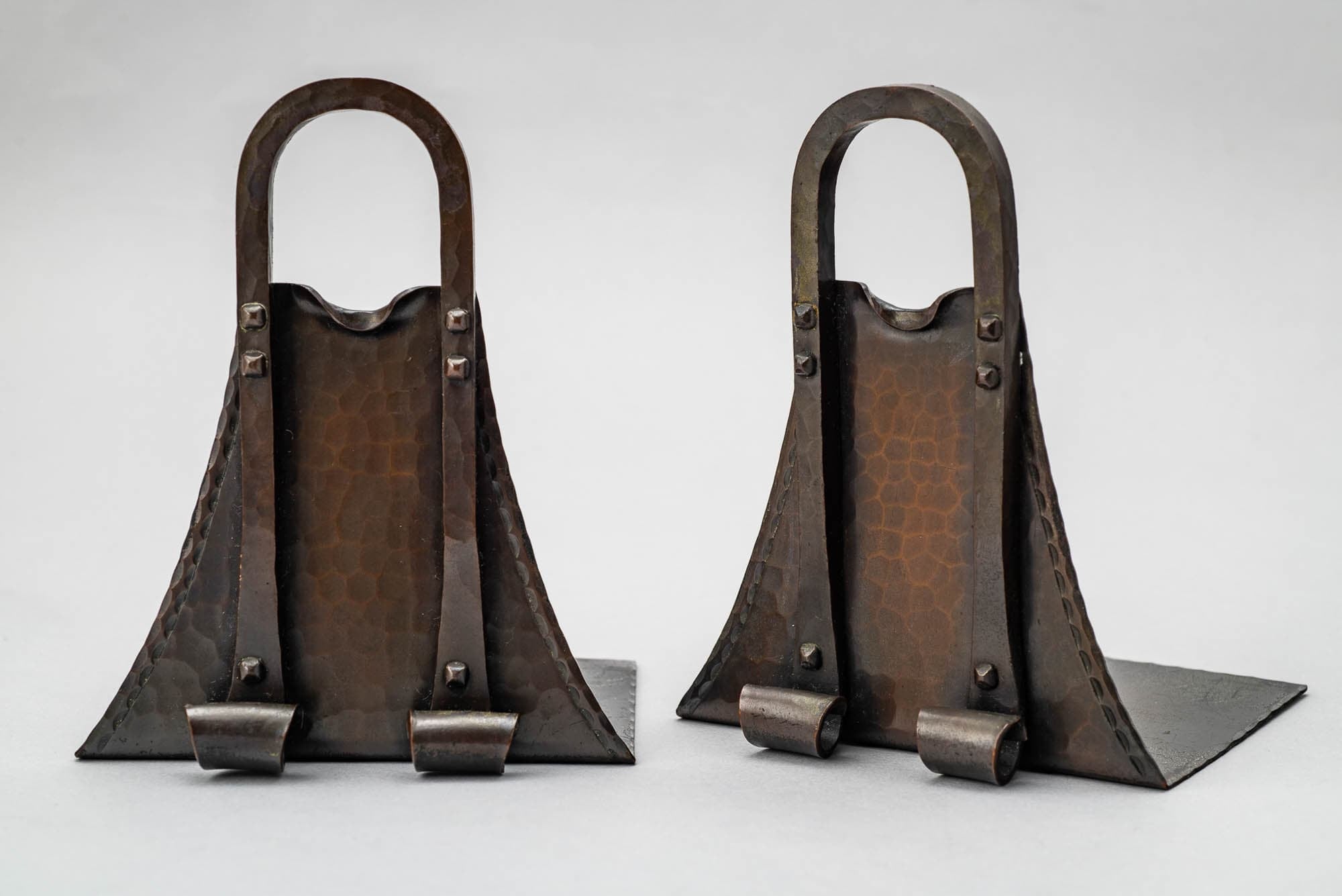 A pair of handcrafted metal bookends with a hammered texture and an arched handle design. The rustic appearance and solid construction suggest they are decorative and functional pieces.