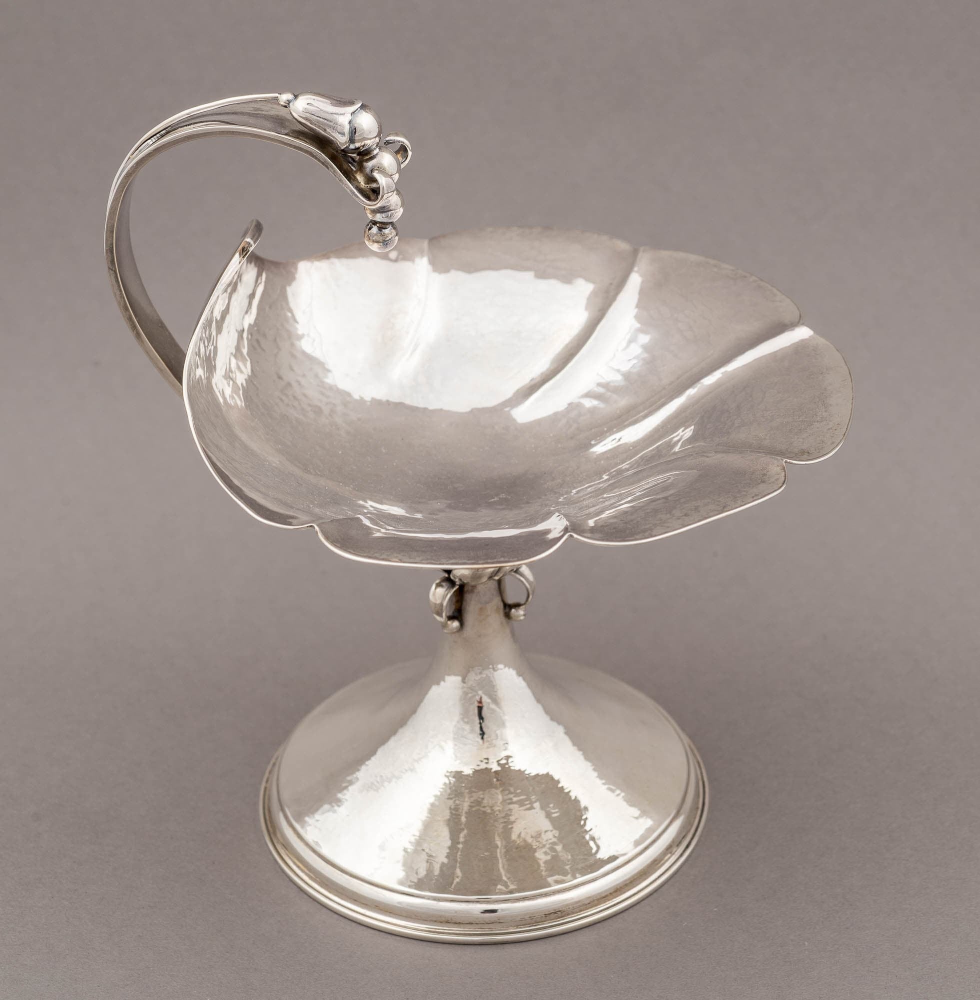 A silver serving dish with a flower petal design and an ornate handle. The polished surface reflects light beautifully, highlighting its elegant craftsmanship.