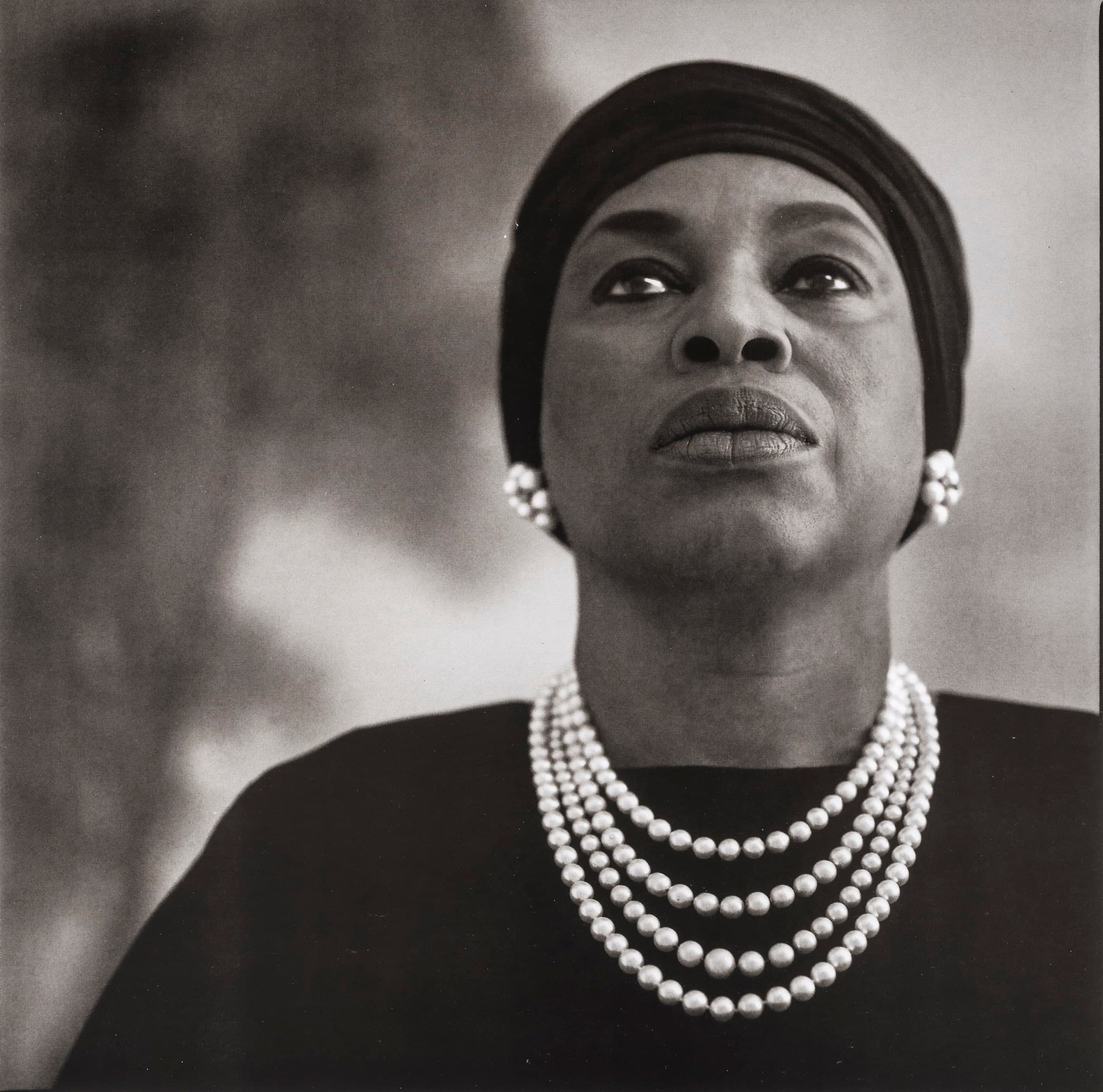 A black and white photo of a woman staring above the camera. She has a prominent pearl necklace on over a black top and pearl earrings. She's wearing a plain black hat. Her gaze is one of power and determination.
