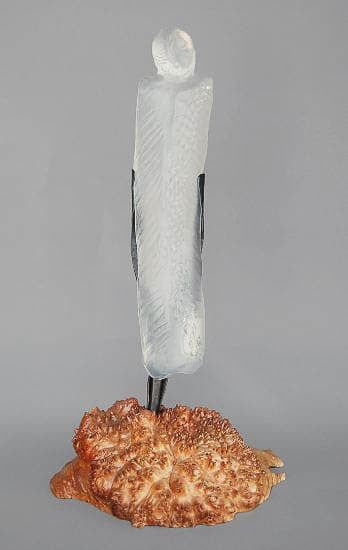 A glass sculpture of a tall, abstract figure with a textured surface, mounted on a rough, natural wooden base. The translucent quality of the glass contrasts with the earthy base.