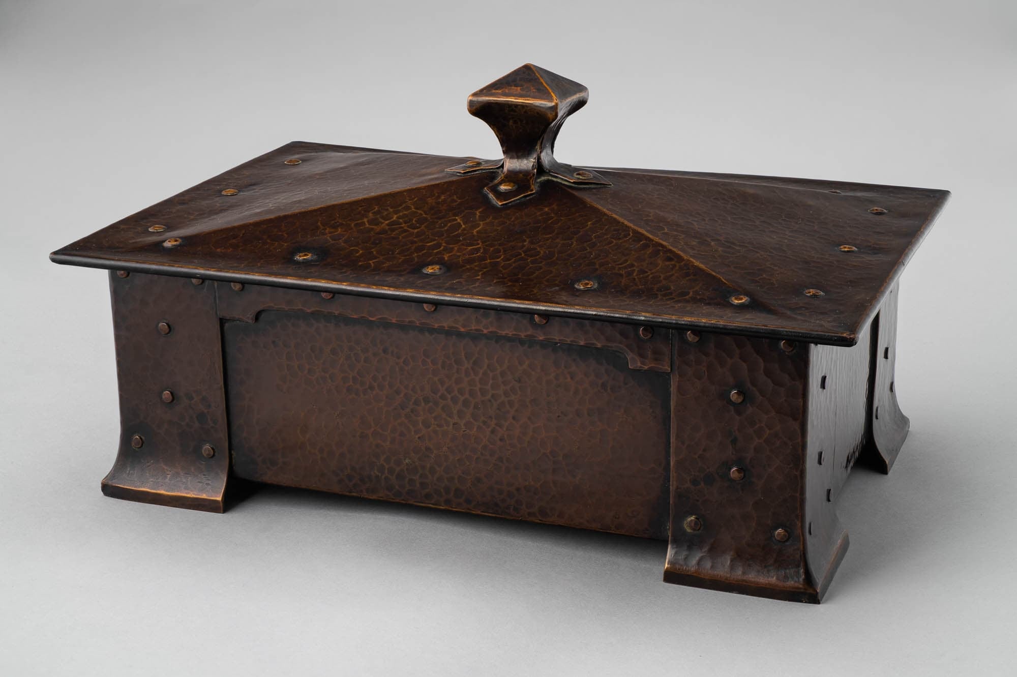 A hammered copper box with a pyramid-shaped lid and decorative rivets. The artisanal craftsmanship and aged patina highlight its functional and decorative purposes.