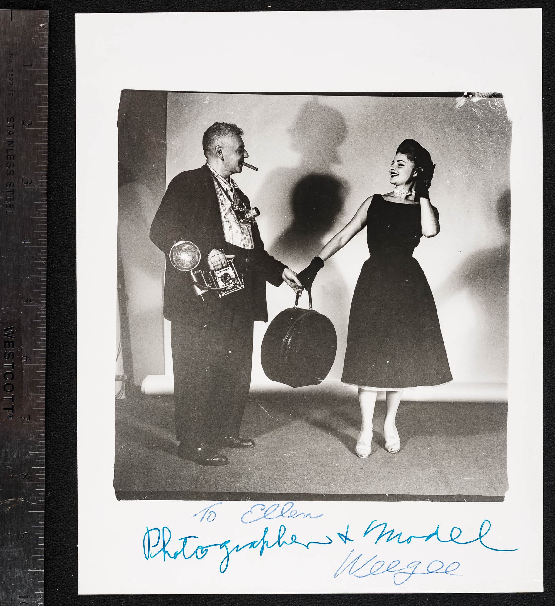 A black-and-white photograph of a photographer and a model. The photographer, holding a camera, smiles at the model, who poses with a suitcase. The image is signed at the bottom.