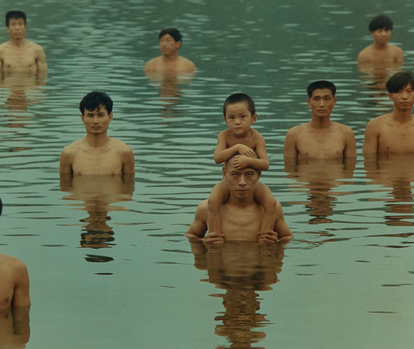 8 shirtless men stand in water up to their chests, staring at the camera. All have black hair and appear to be of Asian descent. One man near the front holds a child on his shoulders whose arms are wrapped around his forehead.