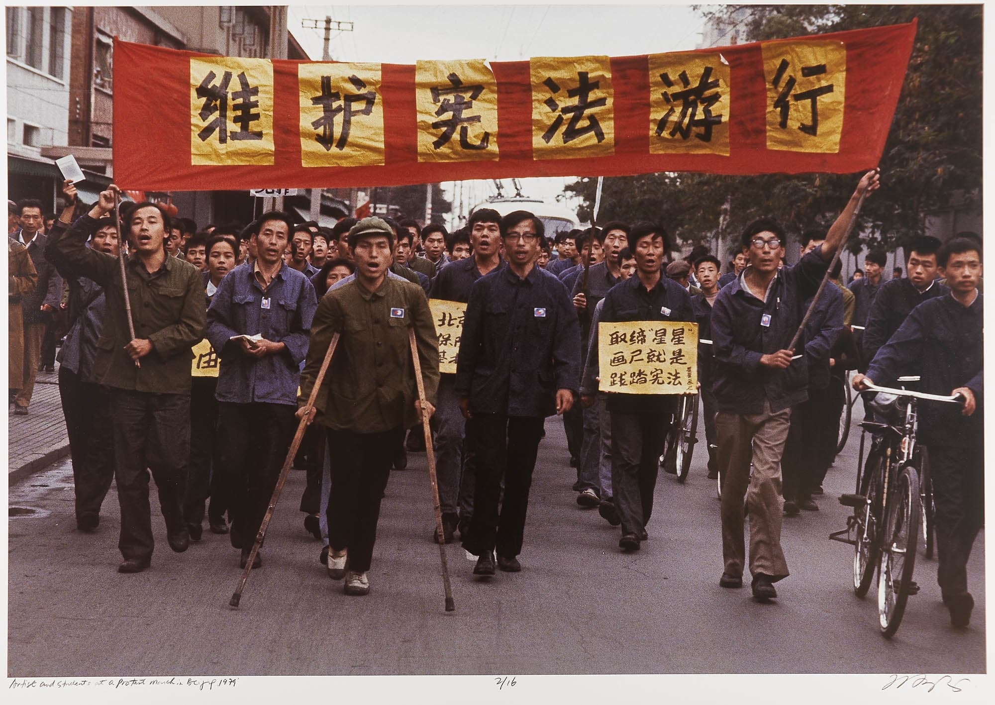 A protest march with people holding signs and a large red and yellow banner. The marchers, including people on crutches, appear determined and unified in their cause.