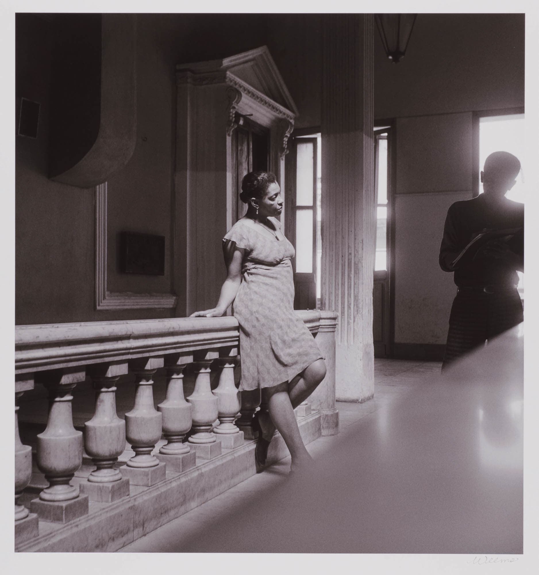 A black and white photograph of a black woman leaning against a balustrade, with a man in the background. The setting is a grand interior space, filled with light and shadow.