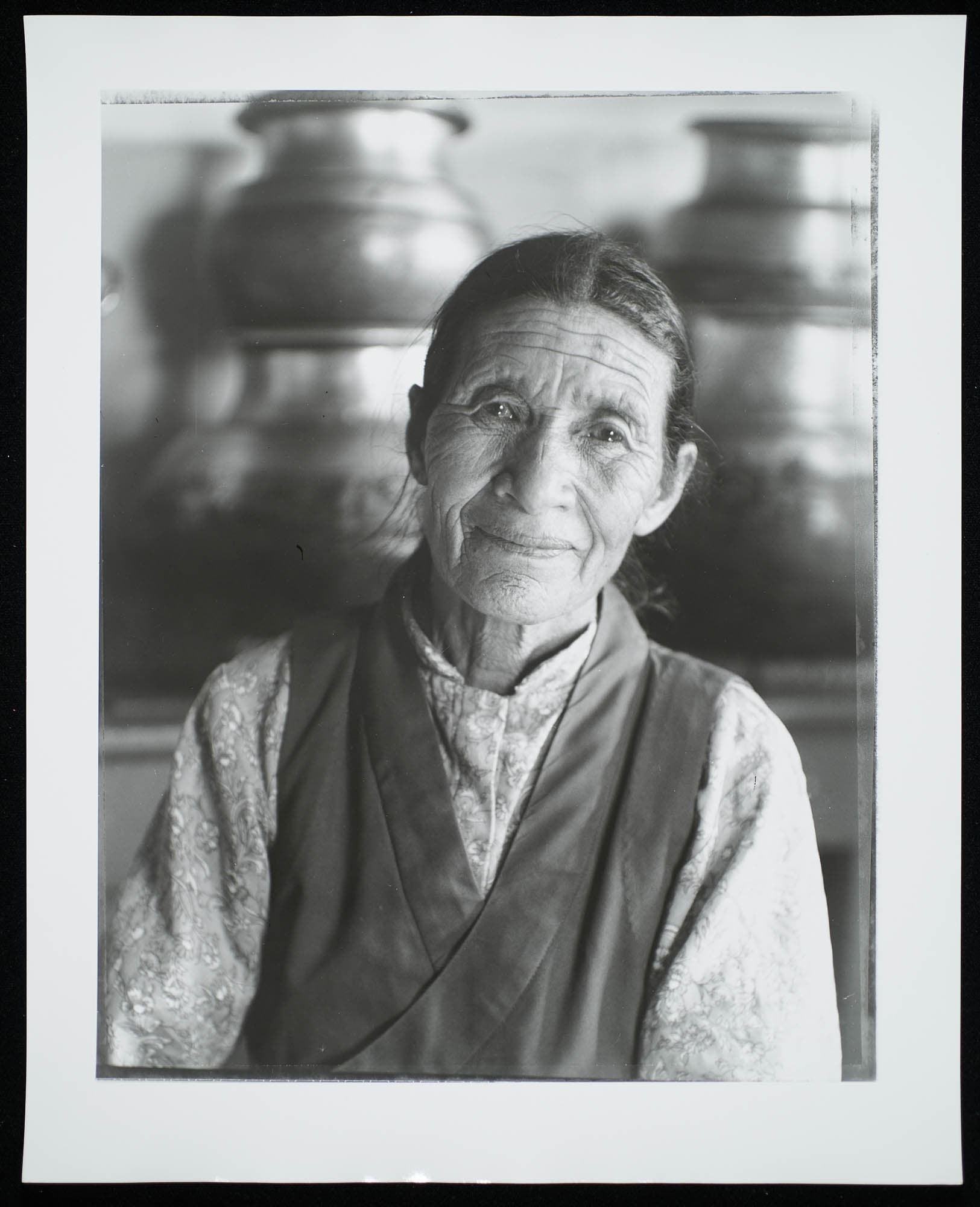 A black-and-white photograph of an elderly woman with a gentle expression, wearing traditional clothing with large pots in the background.