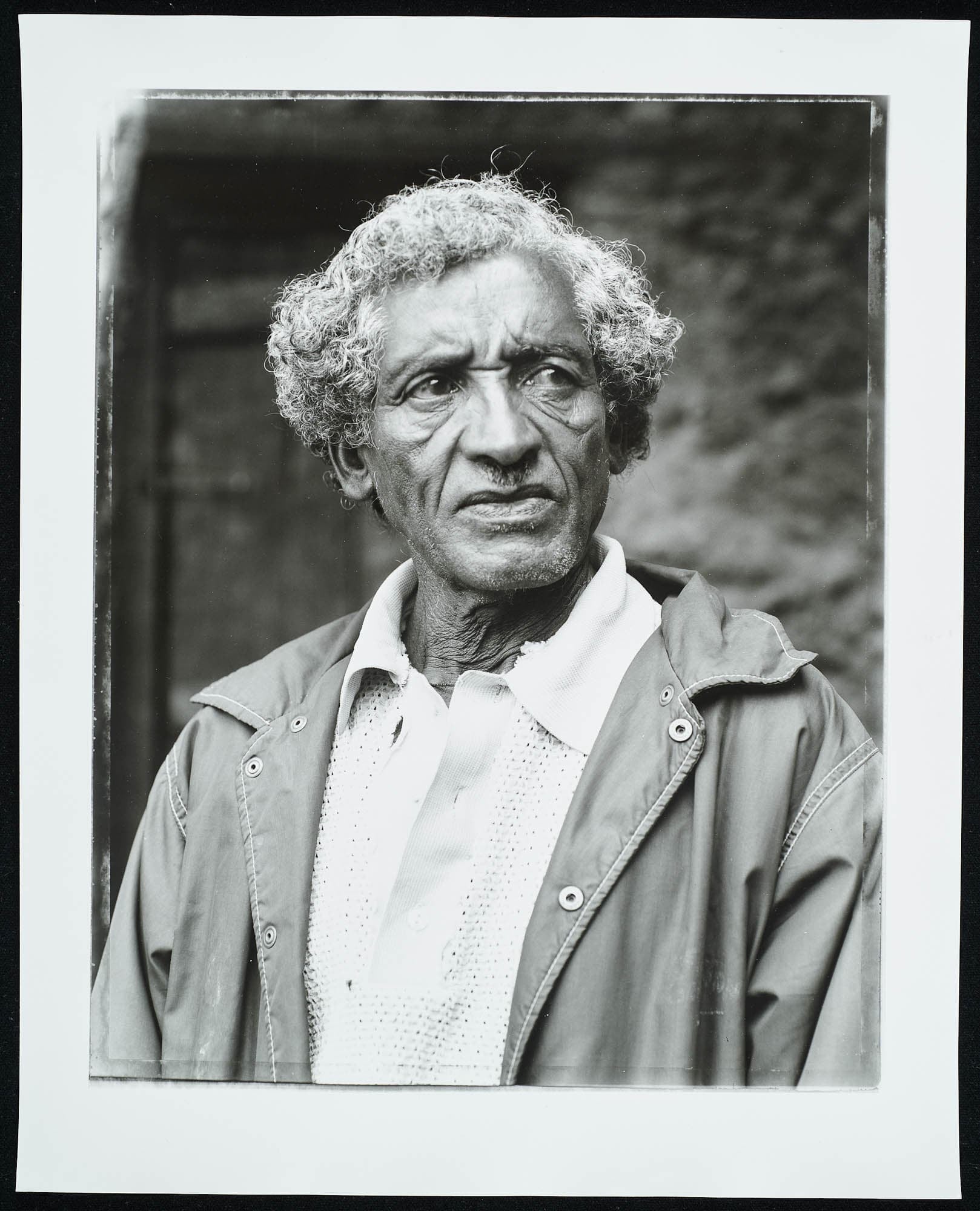 A black-and-white photograph of an elderly man with a serious expression, wearing a light-colored shirt and a jacket, standing against a blurred background.
