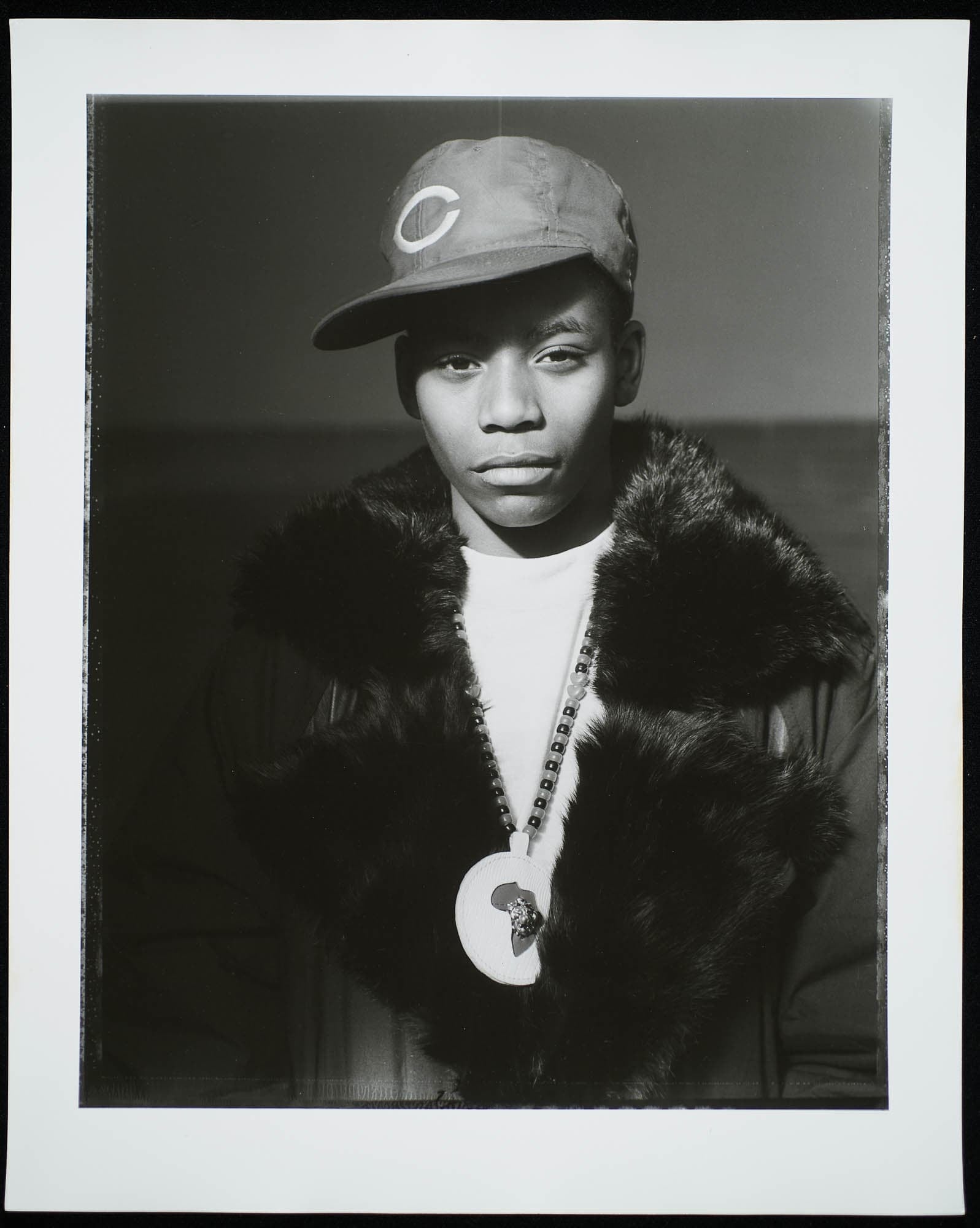 A black-and-white photograph of a young person wearing a baseball cap and a fur-lined jacket, with a medallion necklace, looking directly at the camera.