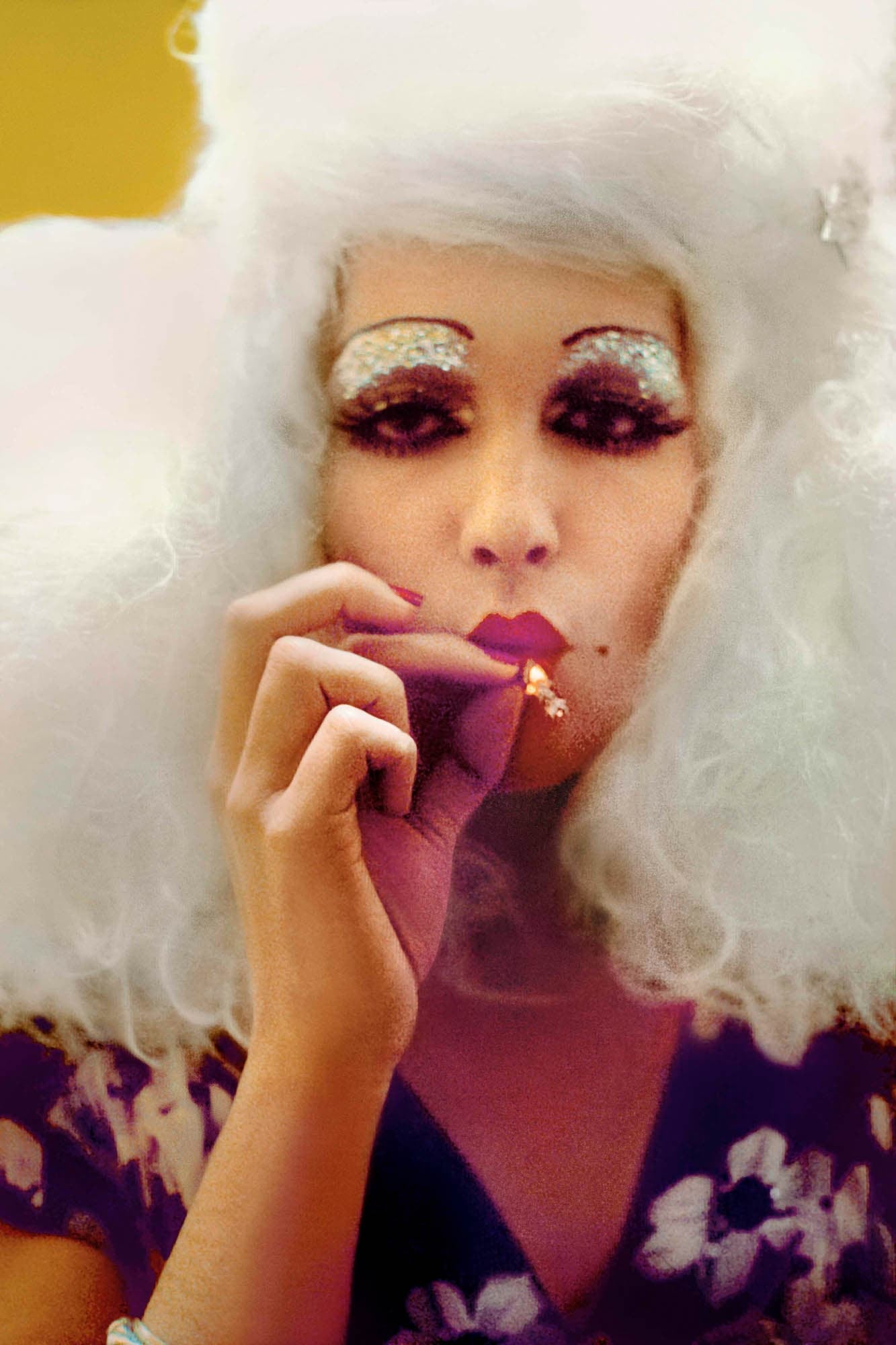 A close-up photograph of a person with a white wig, dramatic eye makeup, and smoking a cigarette. The person has a mysterious and glamorous appearance.