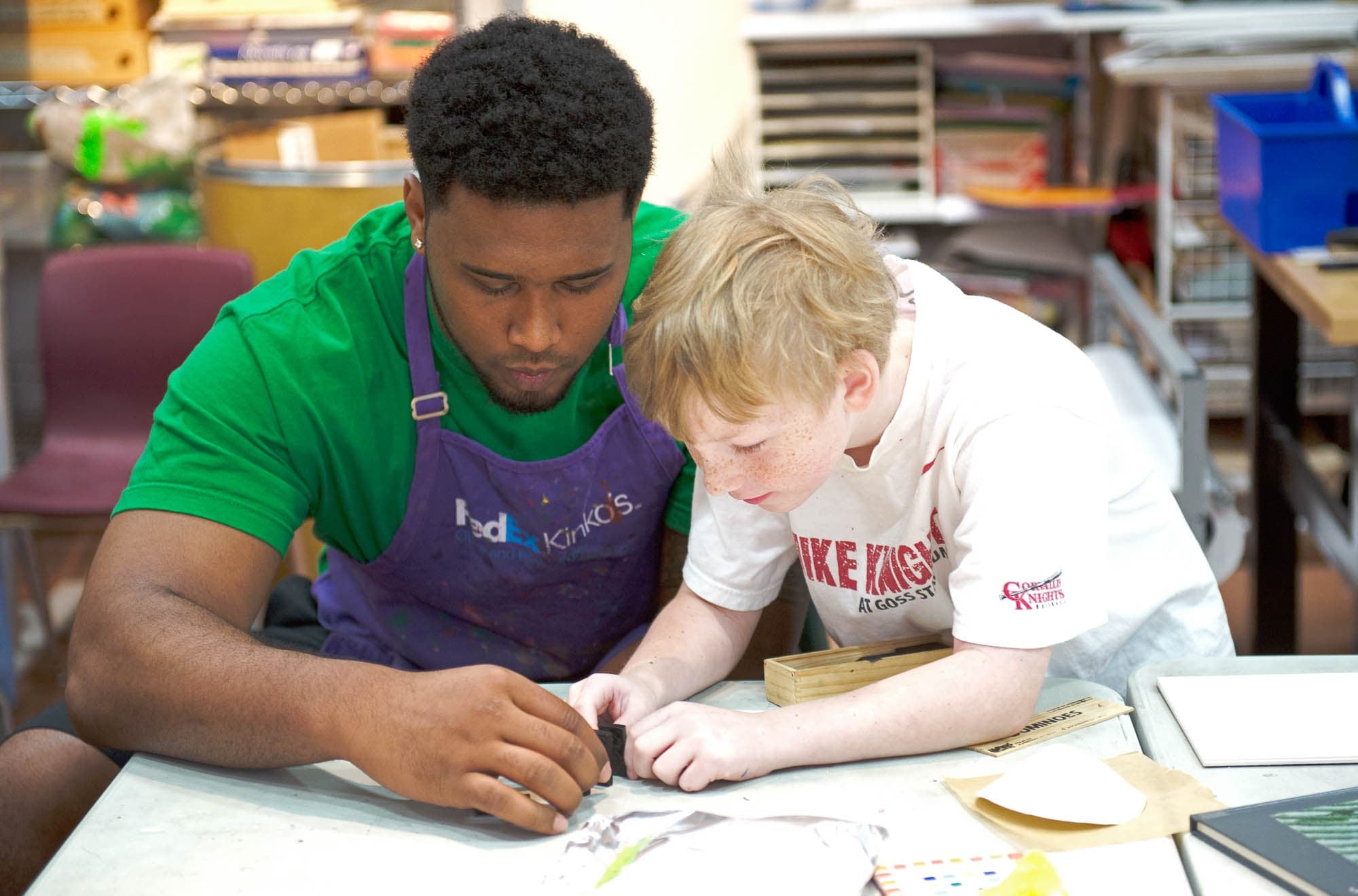 A mentor and a child are engaged in a hands-on activity at a table. The mentor is wearing a green shirt and a purple apron, while the child is in a white t-shirt. Both are focused on the task, fostering a collaborative learning environment.