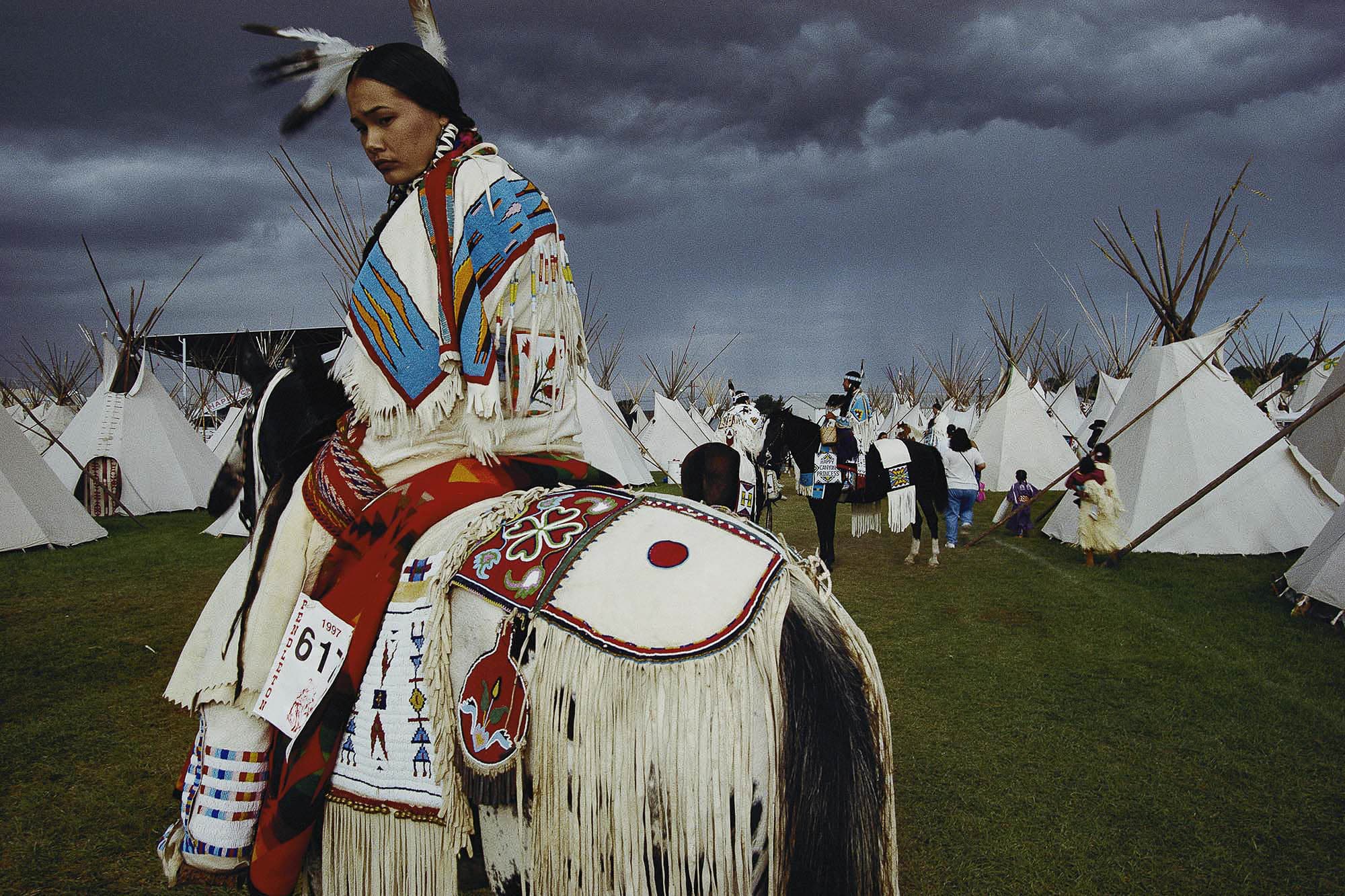 A photograph of a Native American woman in traditional attire riding a horse at a gathering with tipis in the background. The sky is dark and ominous, creating a striking contrast with the colorful garments.