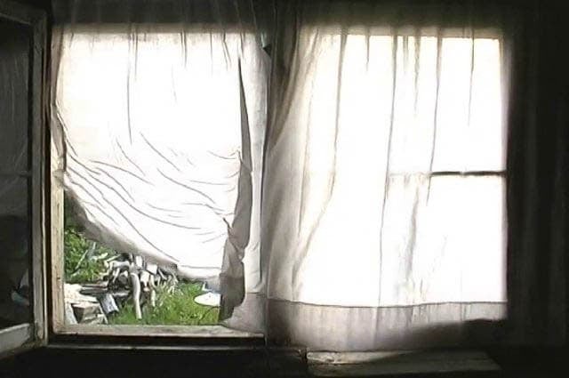 A photograph of a window with sheer curtains blowing in the wind, revealing a glimpse of an outdoor scene with scattered items.