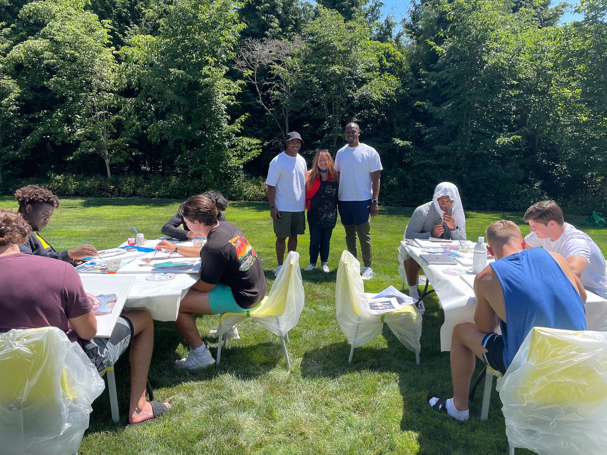 A group of young people sitting at tables outdoors, engaged in an art workshop. Two mentors stand behind them, overseeing the activity. The setting is lush and green, suggesting a creative and inspiring environment.