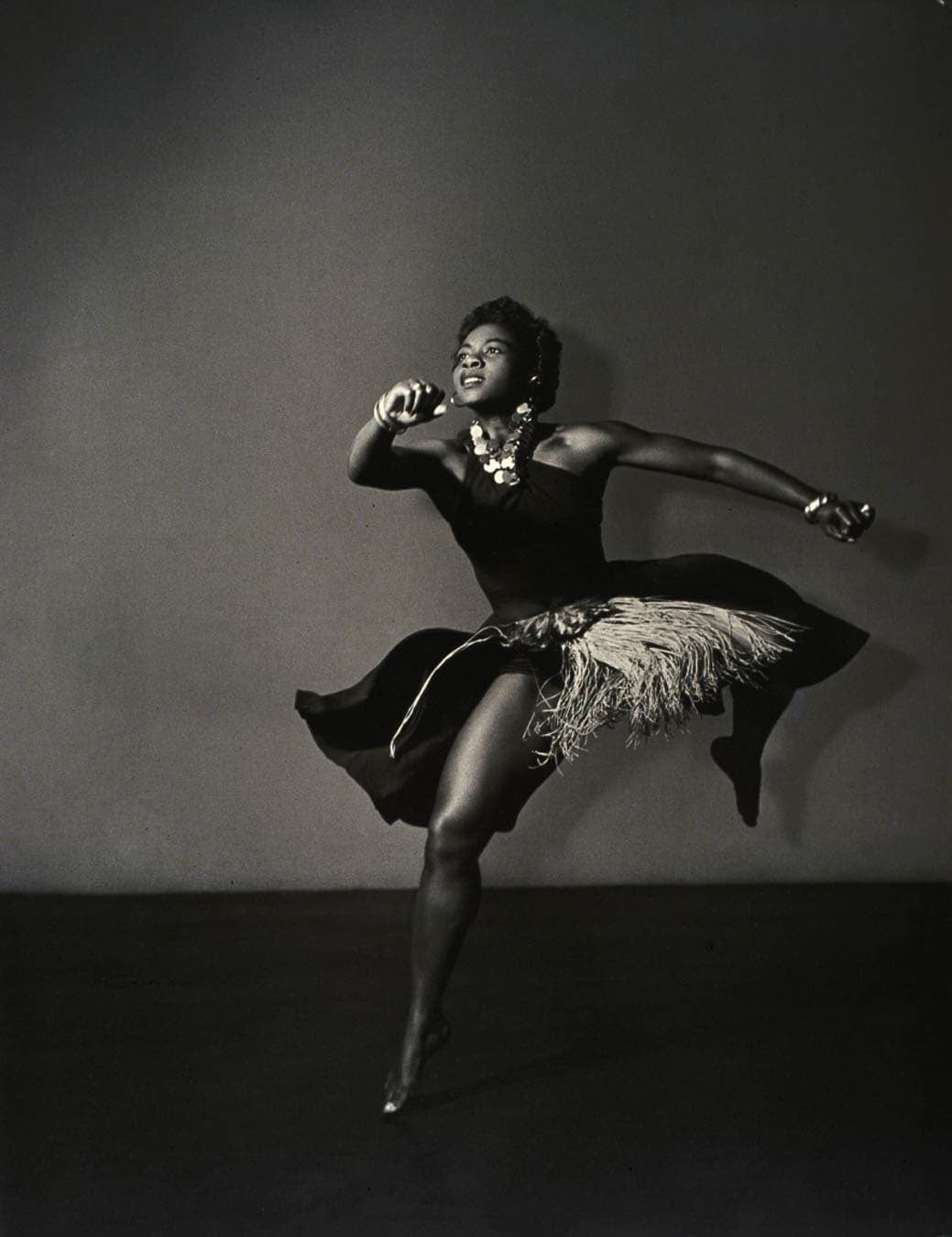 A black and white photograph of a woman dancing energetically, wearing a dress with a feathered skirt.