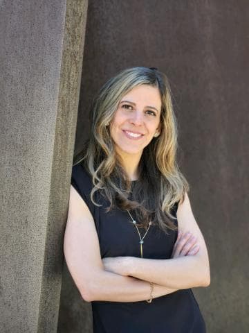 A woman with long wavy hair gives a friendly smile to the camera as she casually leans against a wall with her arms folded across her chest