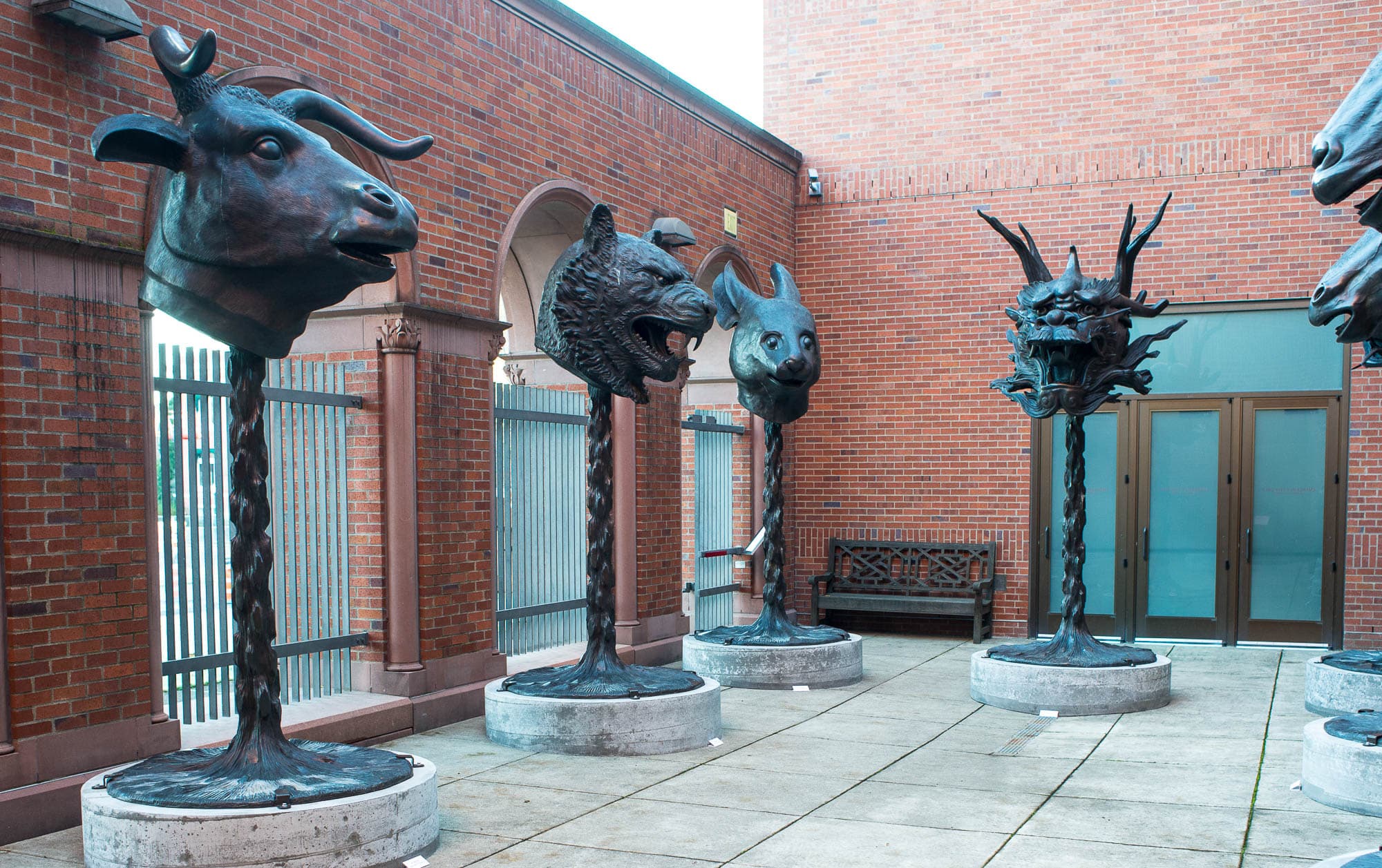 A courtyard installation featuring large bronze animal head sculptures on tall plinths, set against a brick building. This view shows different animal heads.