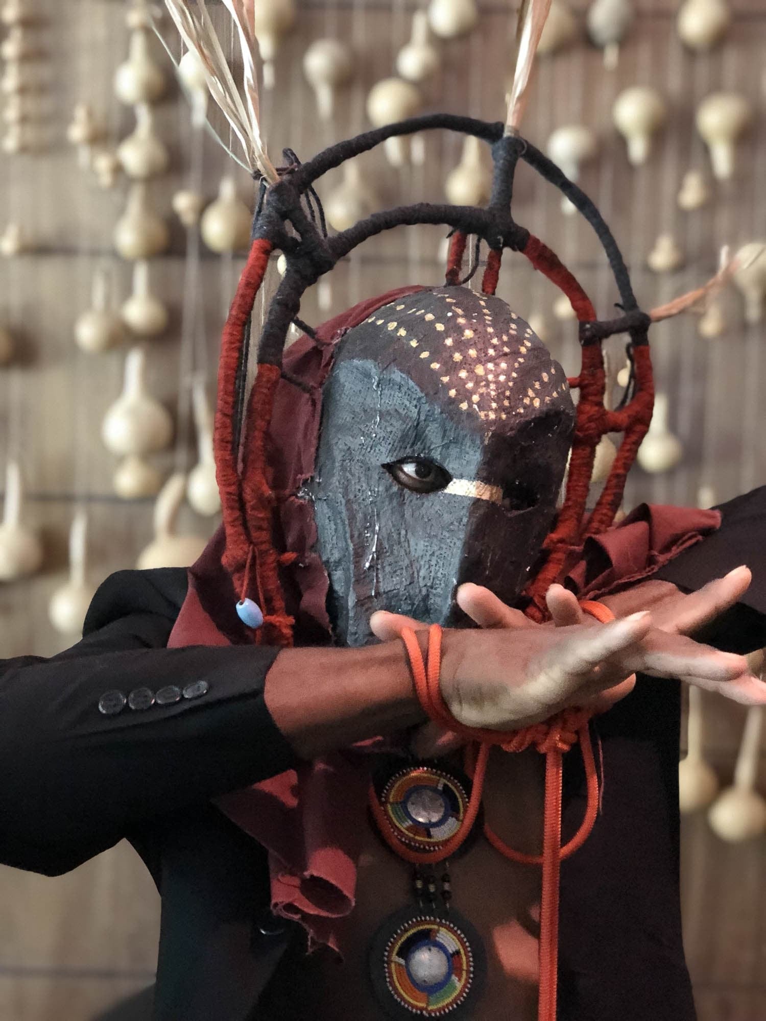 Person wearing a traditional African mask and attire, with red ropes and beadwork, performing a gesture with their hands.