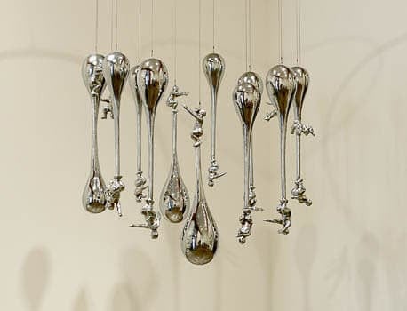 Sculpture featuring multiple chrome droplets hanging from strings, with small human figures climbing on them.