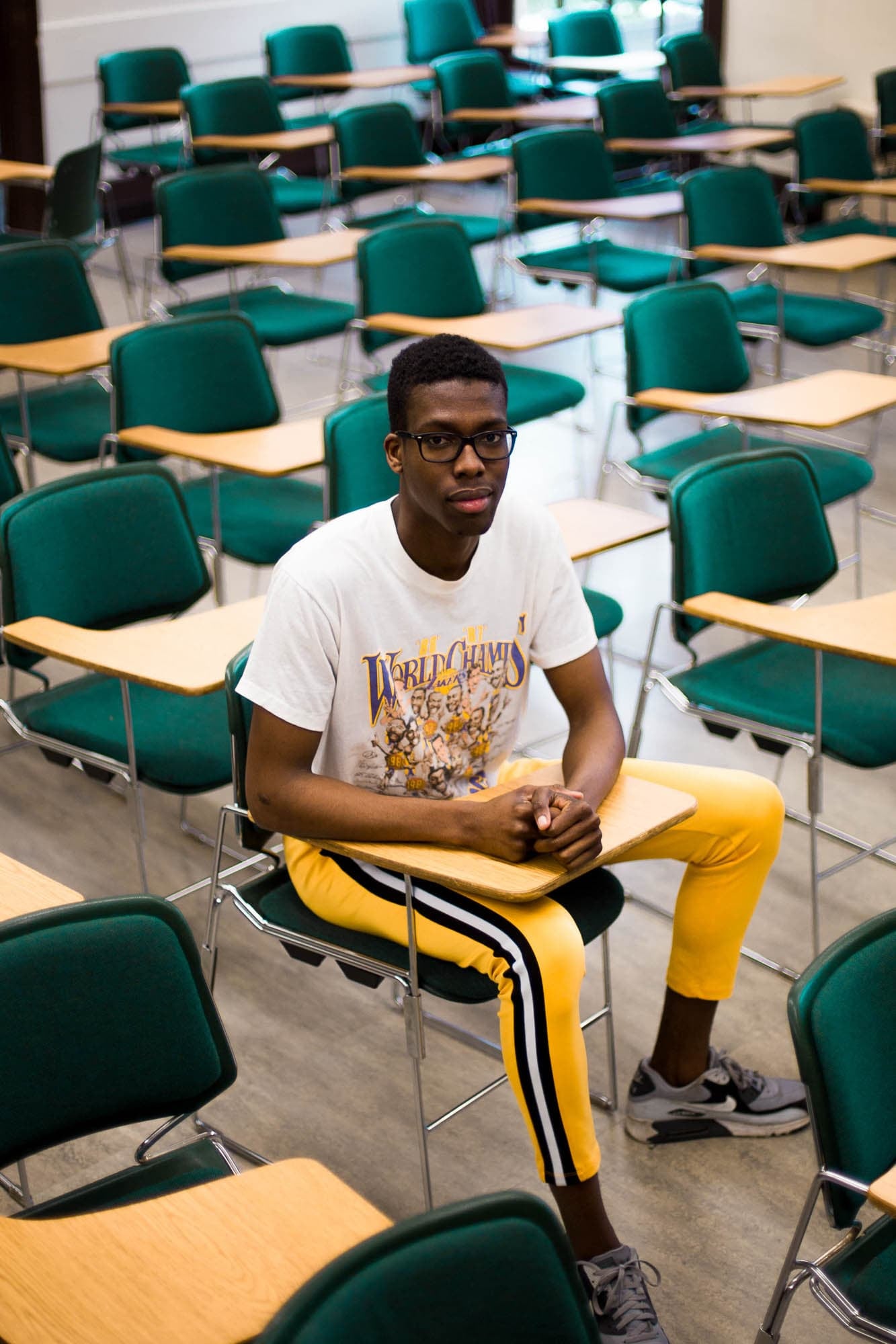 Young man wearing a white t-shirt and yellow pants, sitting in a classroom filled with empty green chairs and desks.