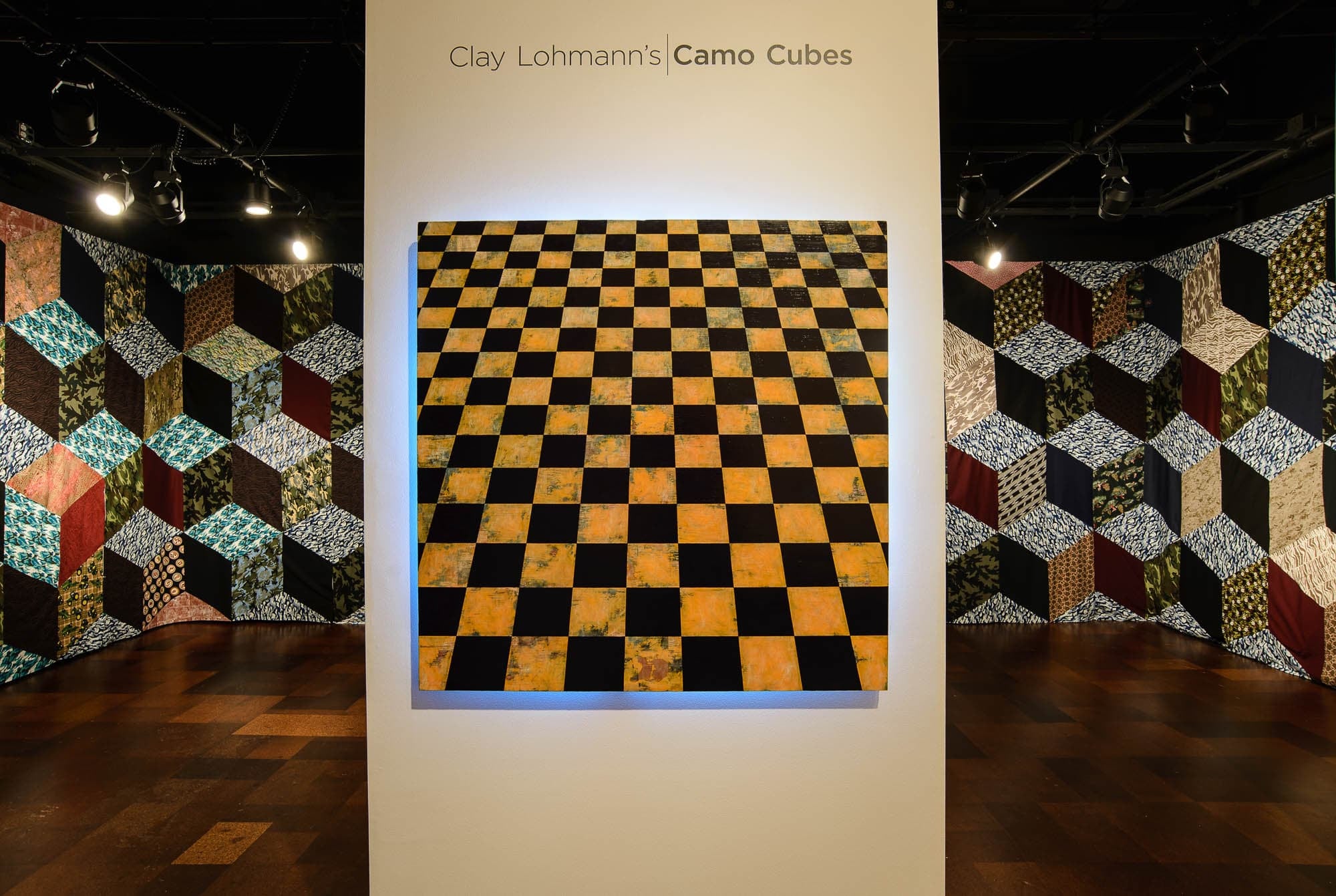 Installation titled 'Clay Lohmannâ€™s Camo Cubes,' featuring a checkerboard pattern and quilt-like wall hangings.