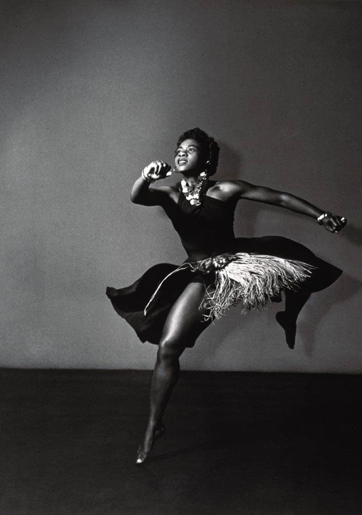 A black and white photograph of a dancer in motion, wearing a dress and adorned with necklaces, captured mid-dance with a graceful pose.