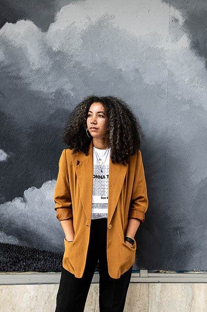 A portrait of Delphine Sims standing against a mural, wearing a tan jacket and a T-shirt, with curly hair framing her face.