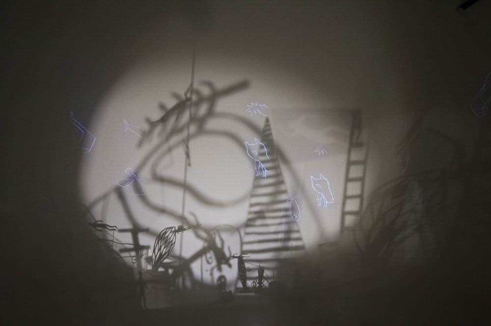 An abstract installation art piece featuring shadowy shapes of ladders, animals, and other objects projected against a dimly lit wall.