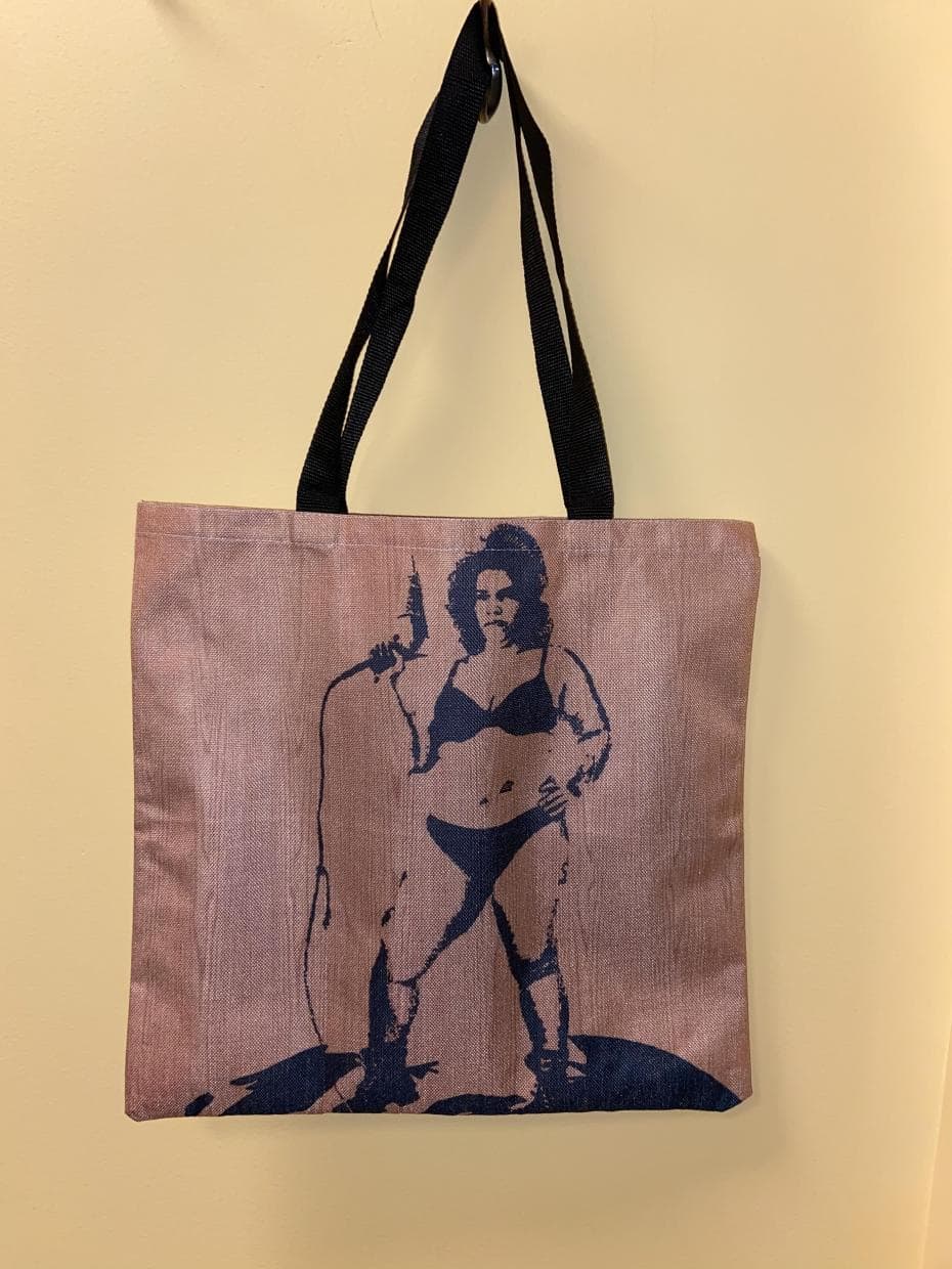 A voluptuous woman with a 1960s beehive hair-do holds a hair dryer cocked up like a gun on this totebag