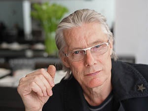 Photograph of a light-skinned person with slicked-back gray hair wearing a wide-collared black jacket and with their hand raised in a loose fist