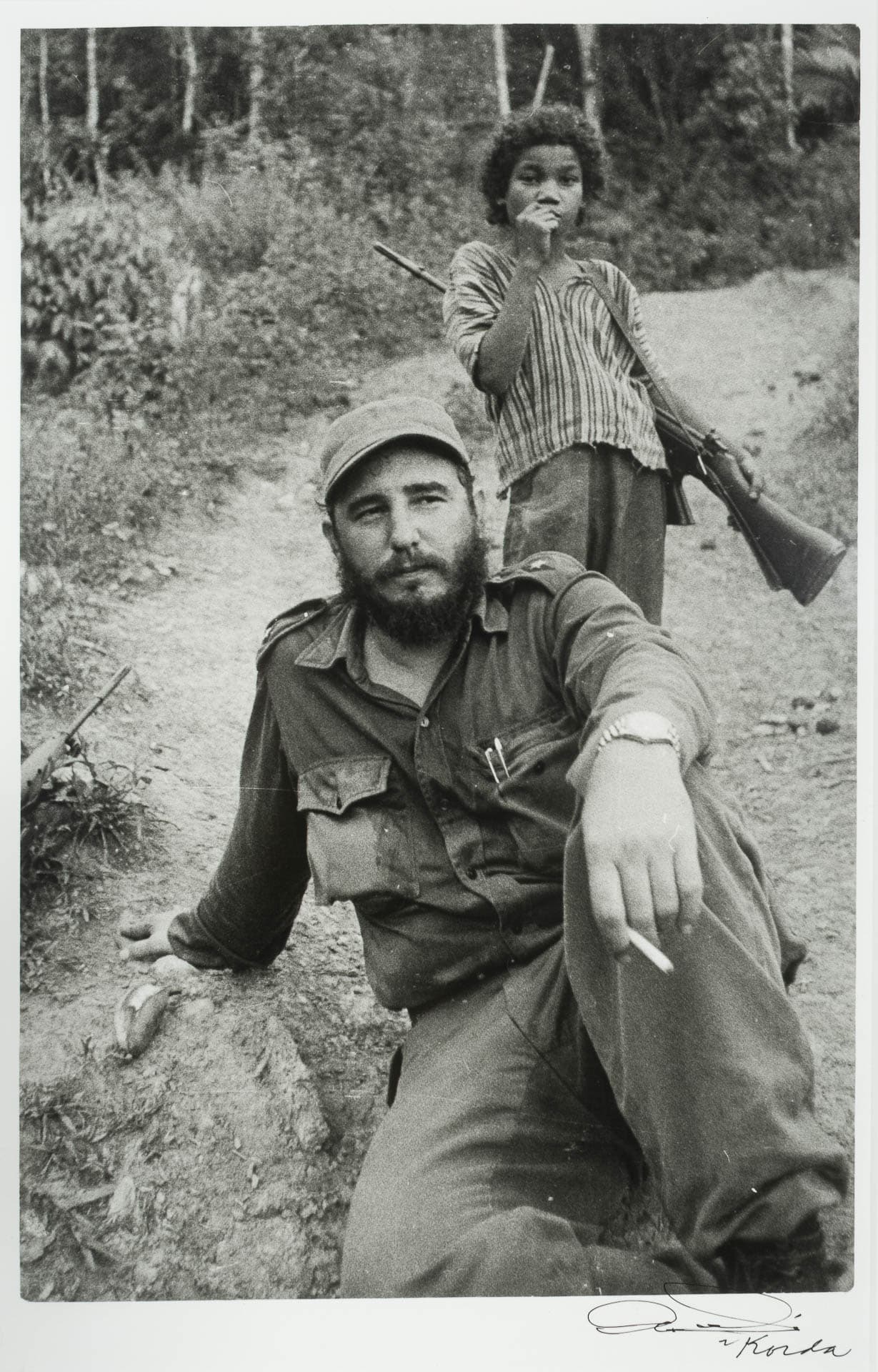 Photograph of a bearded man sitting on a path with a cigarette in his hand and with a child standing behind him with a gun slung over their shoulder