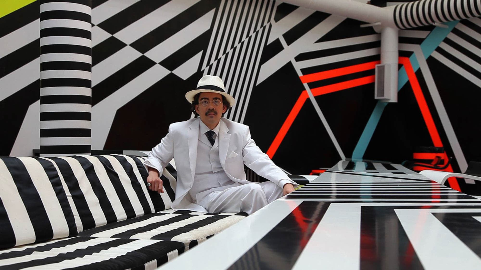 Photograph of a medium-light skinned person sitting on a black and white striped bench wearing a white three-piece suit with a black tie and a black and white fedora. The table and room have black and white stripes in varied directions with one strip each in neon yellow, neon blue, and neon orange.