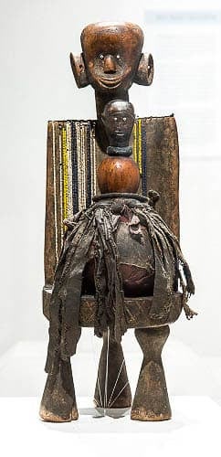 Three-legged wooden chair with a person’s head carved on top and a smaller wooden sculpture shaped like a snowman with fabric strips cinched between the bottom and second spheres balanced on the chair’s seat