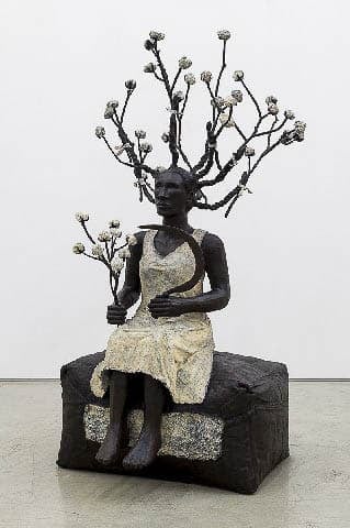Bronze sculpture of a seated woman in a white dress holding a scythe and a branch of cotton. There are cotton branches attached to each of her upright braids.