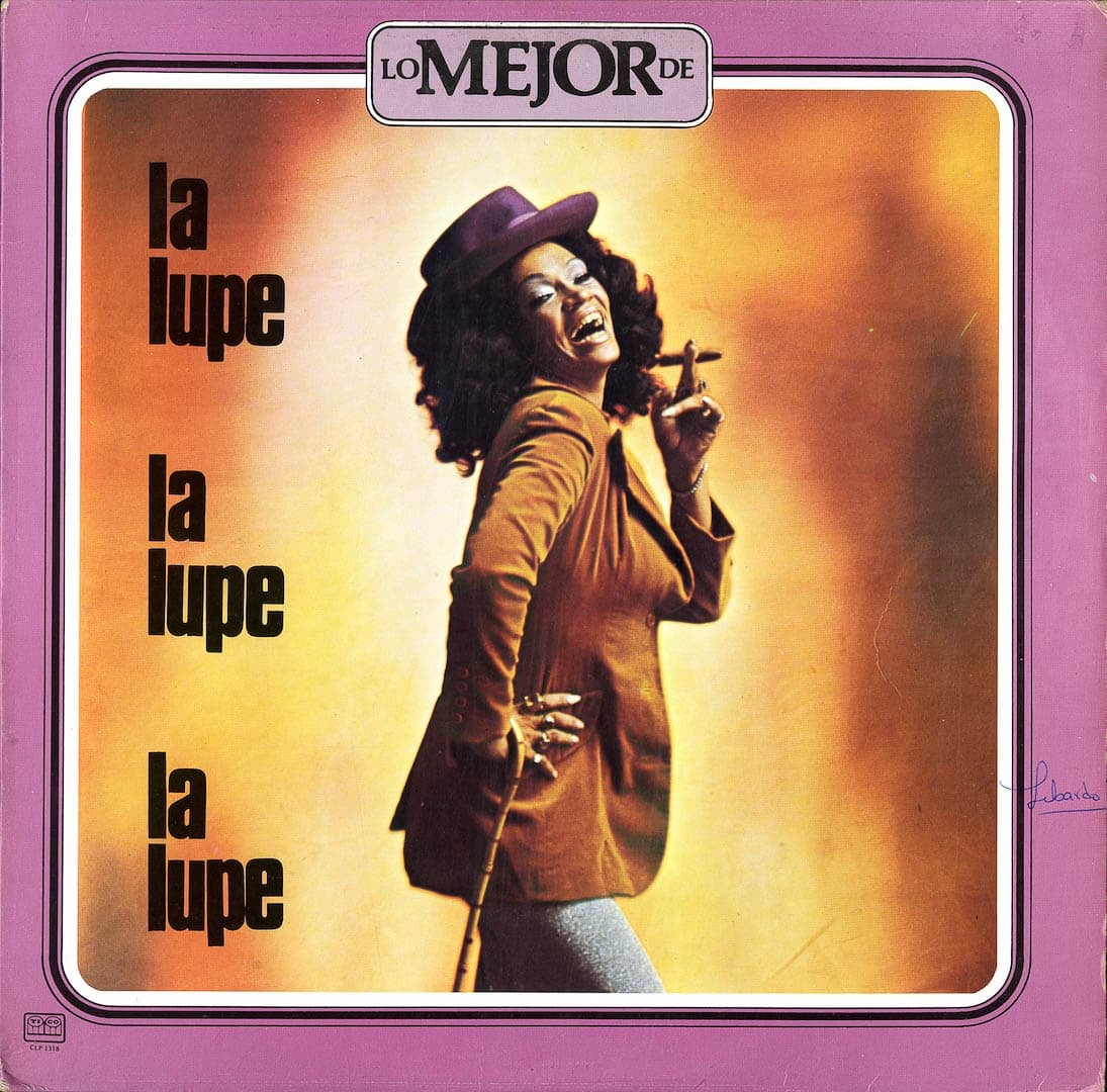Album over for “Lo Mejor de la lupe.” A medium-dark skinned person stands on the cover with one hand on their hip and the other holding a cigar. Their smiling mouth is open and their head is tilted back, surrounded by shoulder-length black curls. They are wearing a plum-colored brimmed hat and a tan jacket. “La lupe” is repeated three times down the left side.