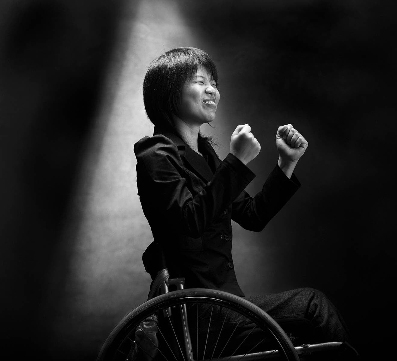 Black and white photo of an Asian person in a wheelchair. Their  clenched fists are raised in front of their chest in either triumph or pain.