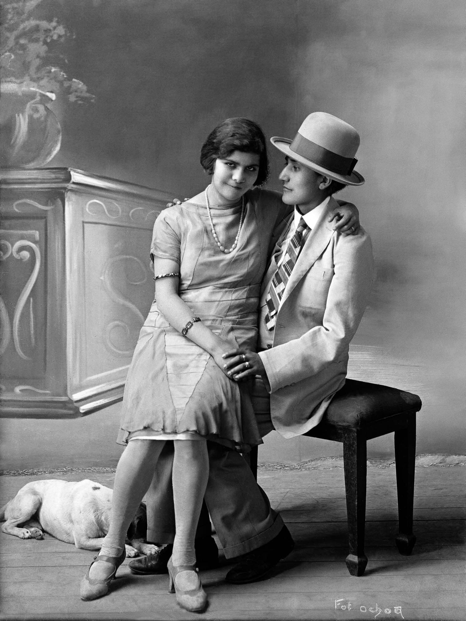 Black and white photograph of a couple sitting on a piano bench with a dog at their feet. One wearing a dress and pearls sits on the lap of the other who wears a suit and hat while they hold hands.