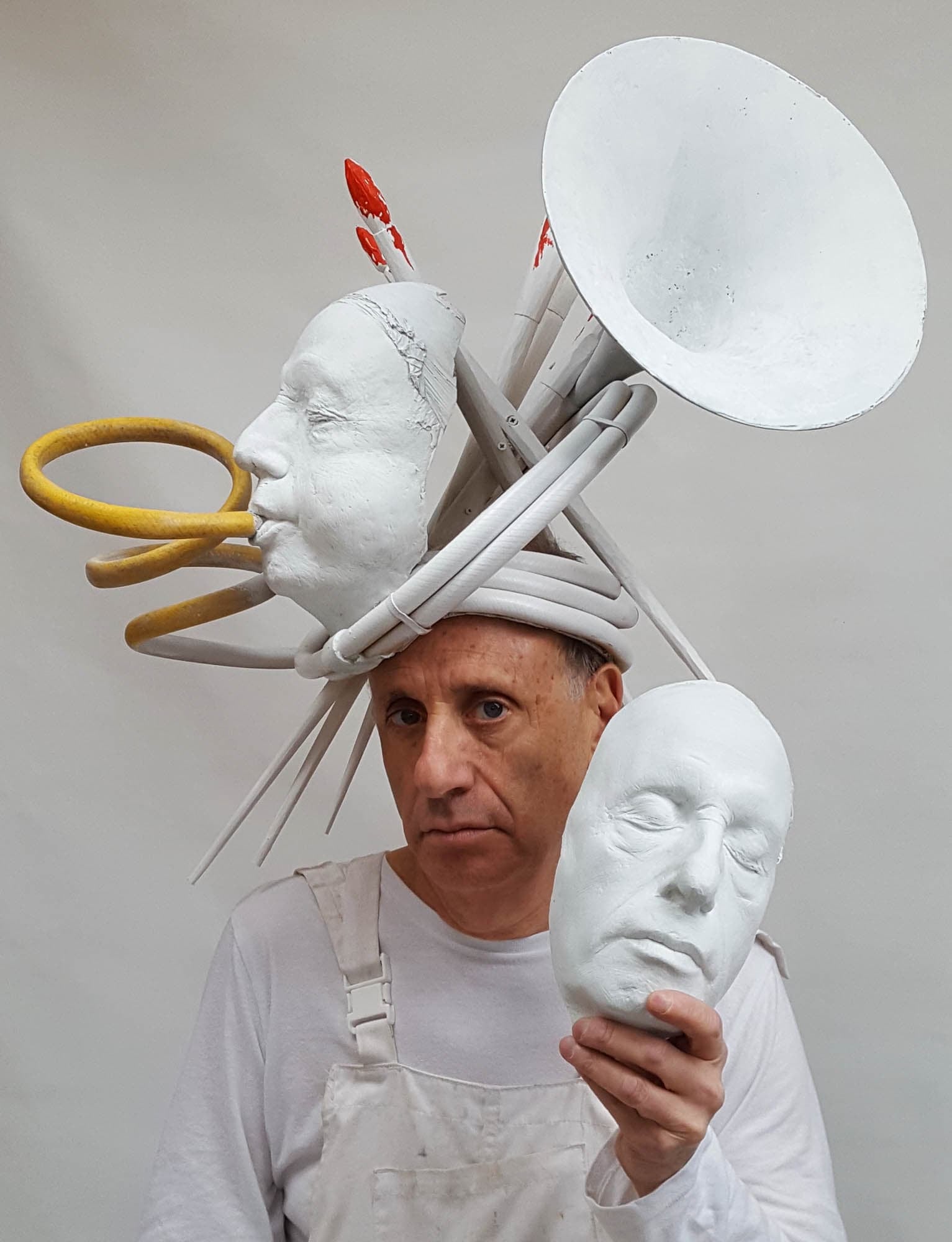 Photograph of Philip Haas, a light-skinned man wearing a long-sleeved white shirt and white coveralls. In his hand is a white plaster death mask. On his head is a white sculpture with a second mask blowing into a Seussian horn interspersed with paintbrushes with red paint on their bristles.
