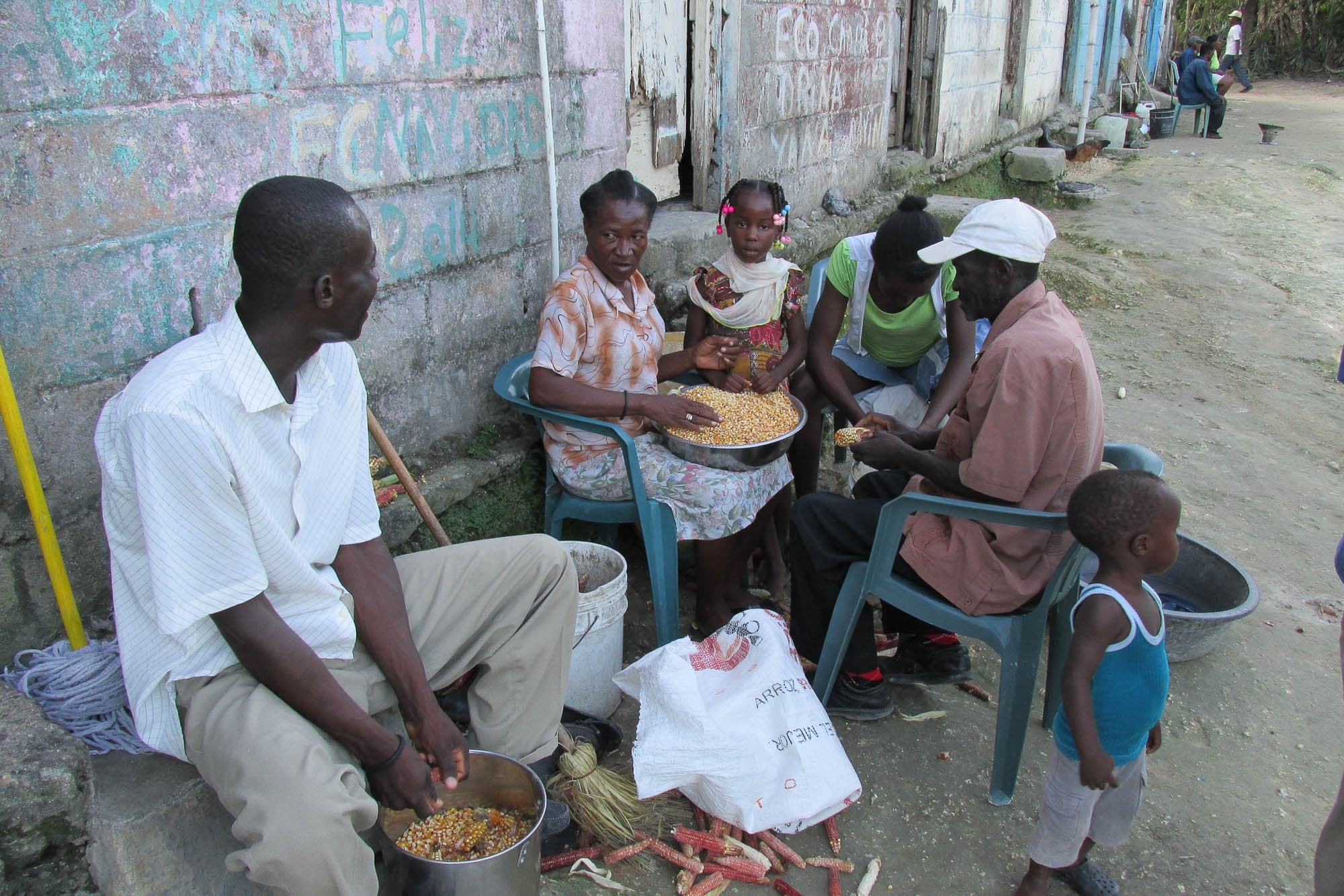 Photograph of a group of dark-skinned people of varying ages sitting outside a cinderblock building shelling corn